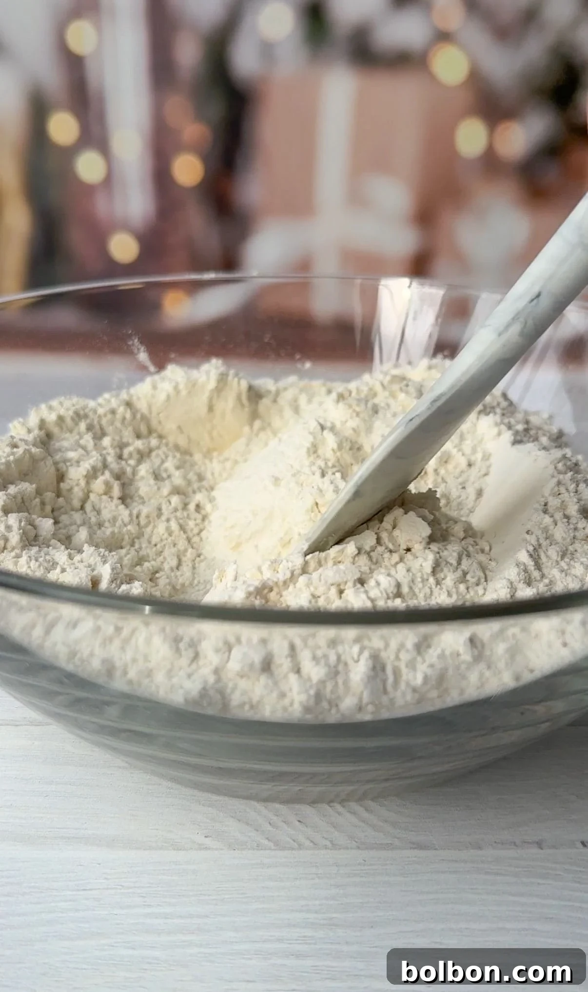A glass bowl filled with flour and a white spatula resting in it. The background is softly blurred with warm, decorative lights and wrapped gifts, creating a festive atmosphere.