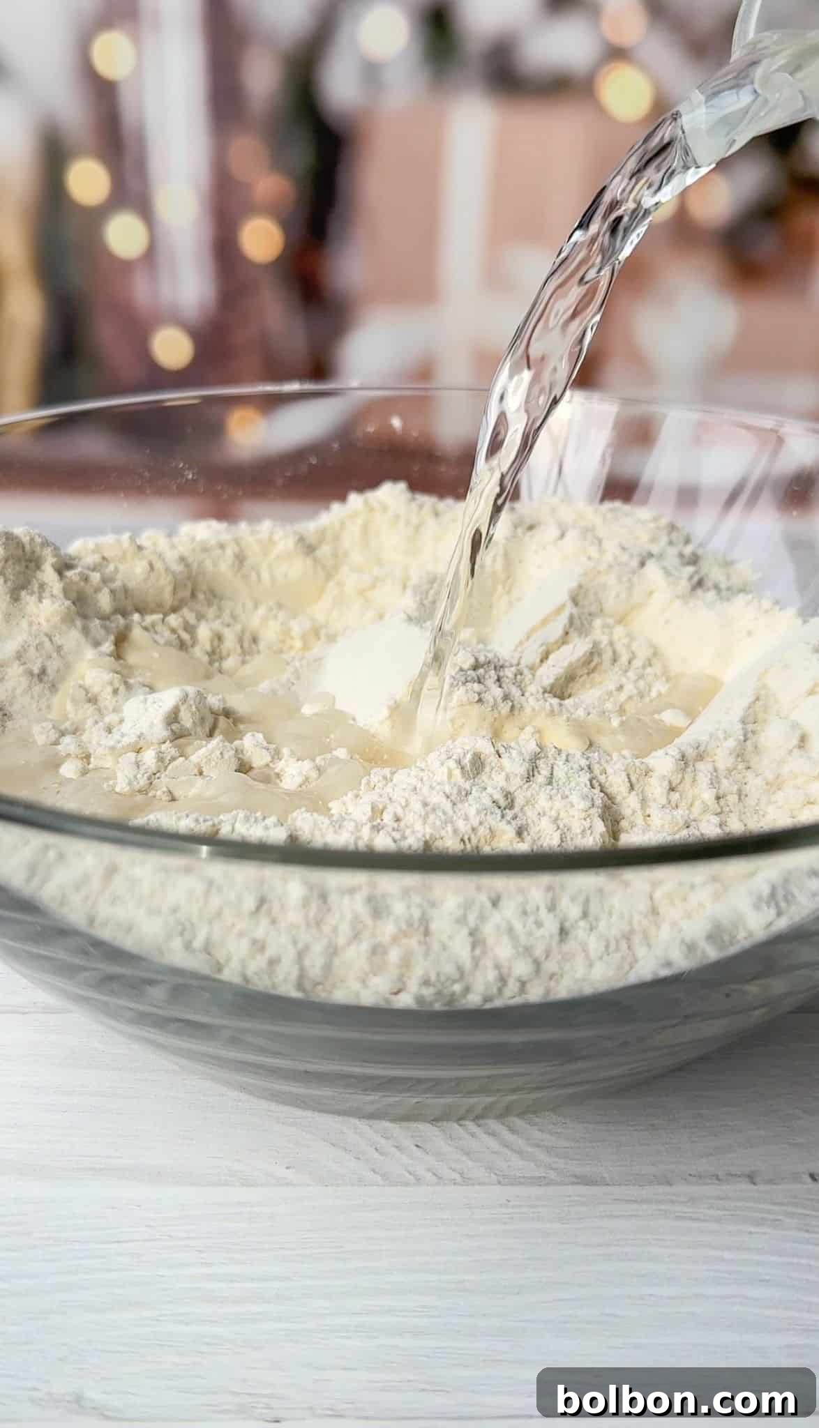 Clear liquid is being poured into a clear glass bowl filled with flour. In the background, there are blurred lights and indistinct shapes, creating a festive atmosphere.