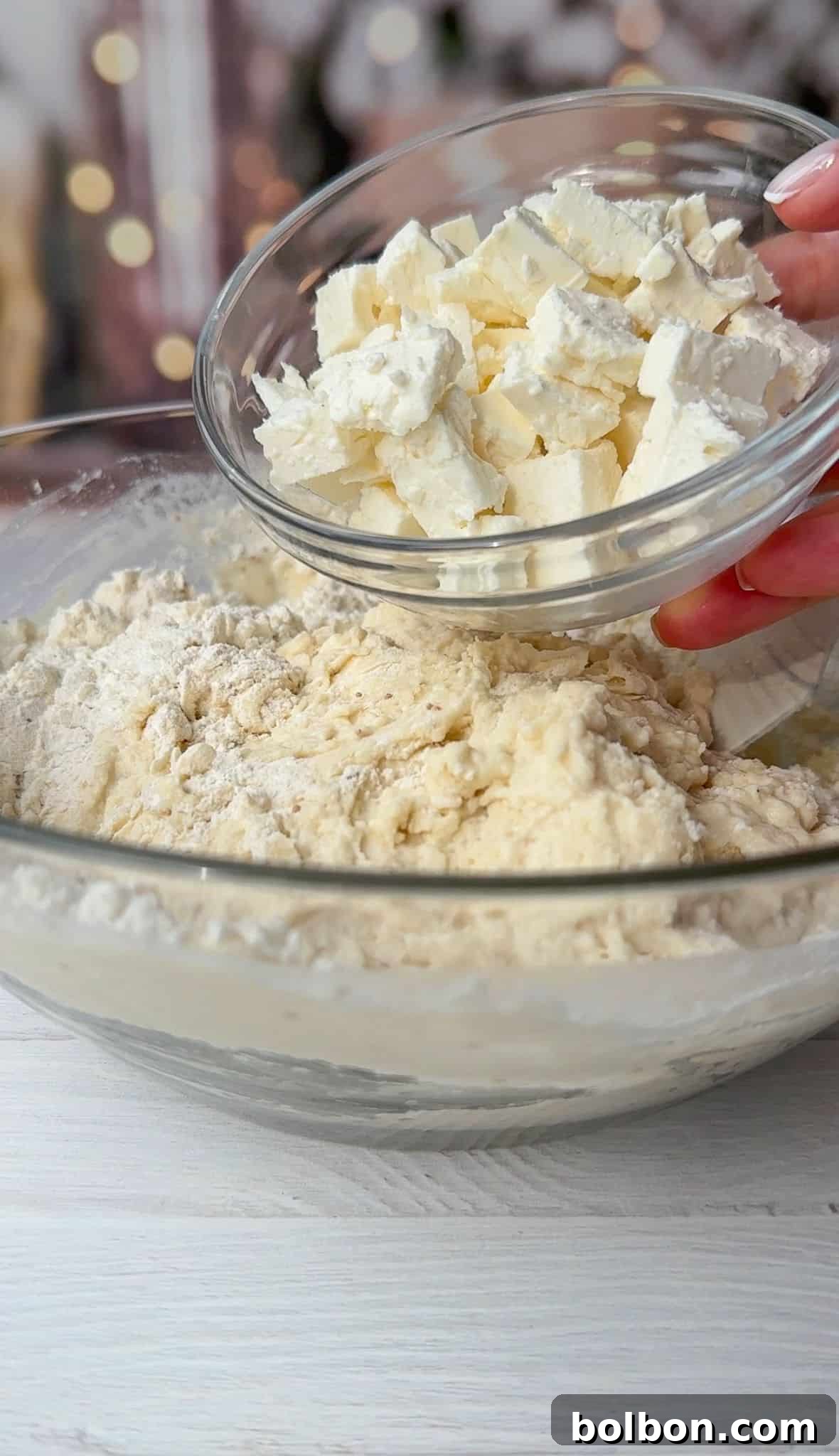 A hand holding a small bowl of cubed butter is adding it to a large mixing bowl filled with flour. The ingredients are on a white wooden surface, with blurred lights in the background.