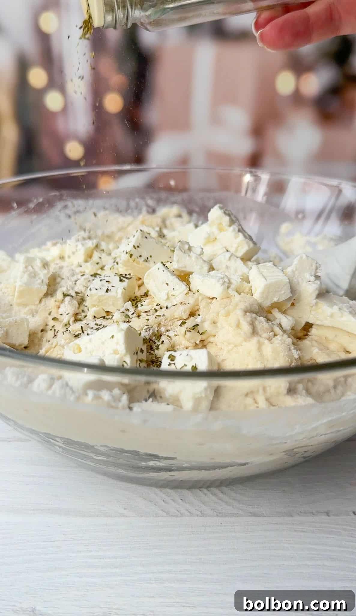 A clear glass bowl filled with chunks of feta cheese, creamy mixture, and herbs being sprinkled from a shaker above. The background is softly blurred, emphasizing the preparation of a dish on a white surface.