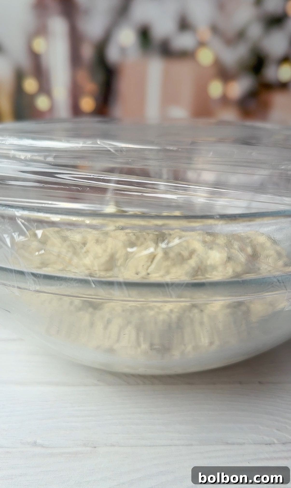 A glass bowl covered with plastic wrap, containing dough, sits on a white surface. Blurred background features soft lighting and hints of holiday decorations.