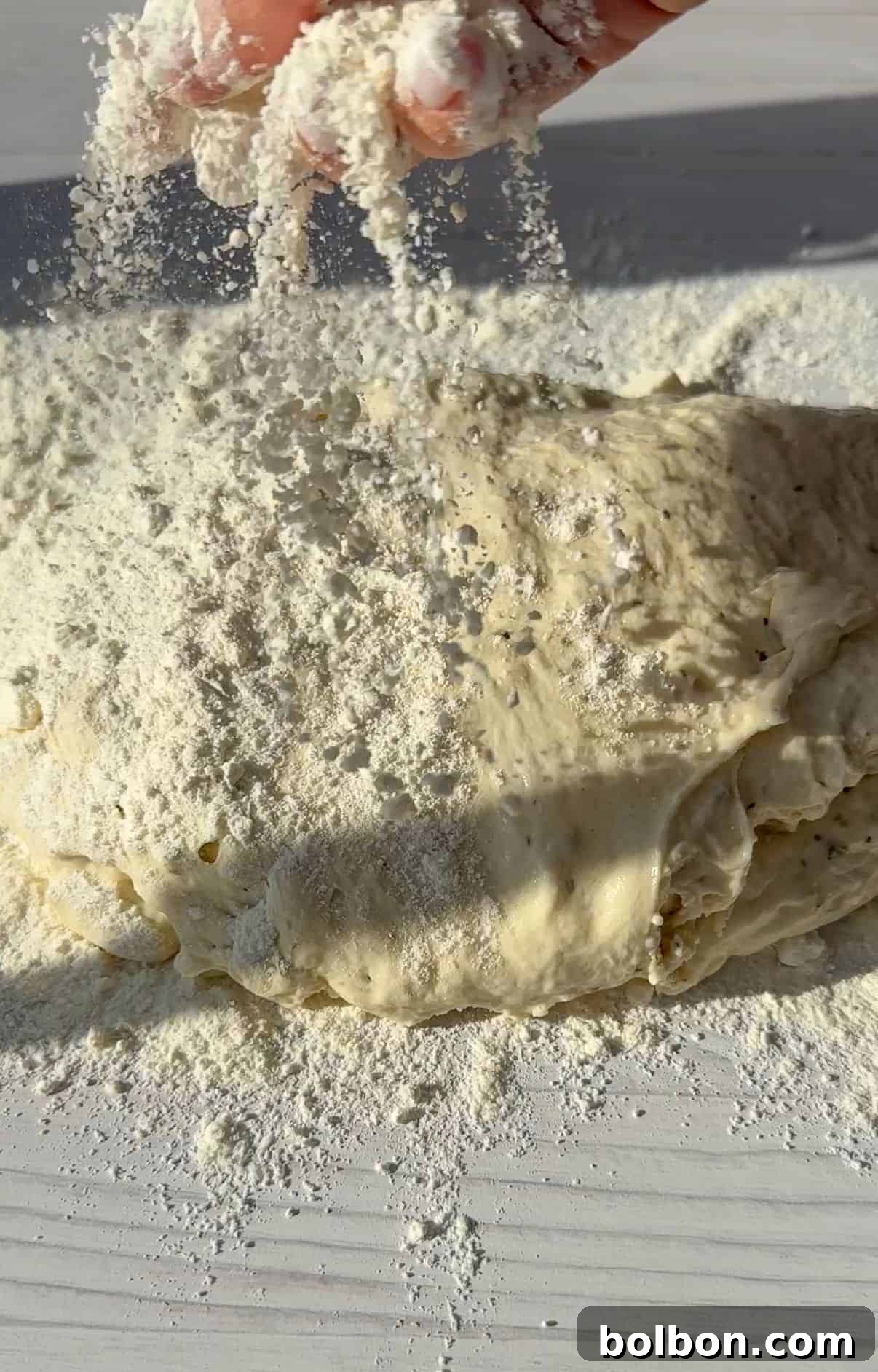 A hand sprinkling flour over a mound of no knead dough on a white surface, with sunlight casting soft shadows.