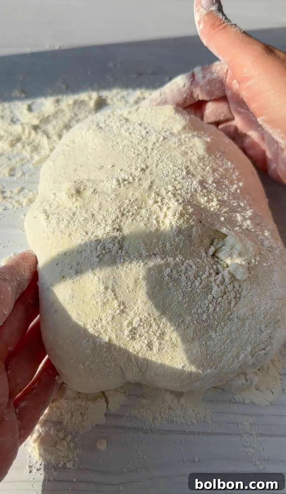 Hands kneading a dough ball covered in flour on a light-colored surface, with sunlight casting shadows.