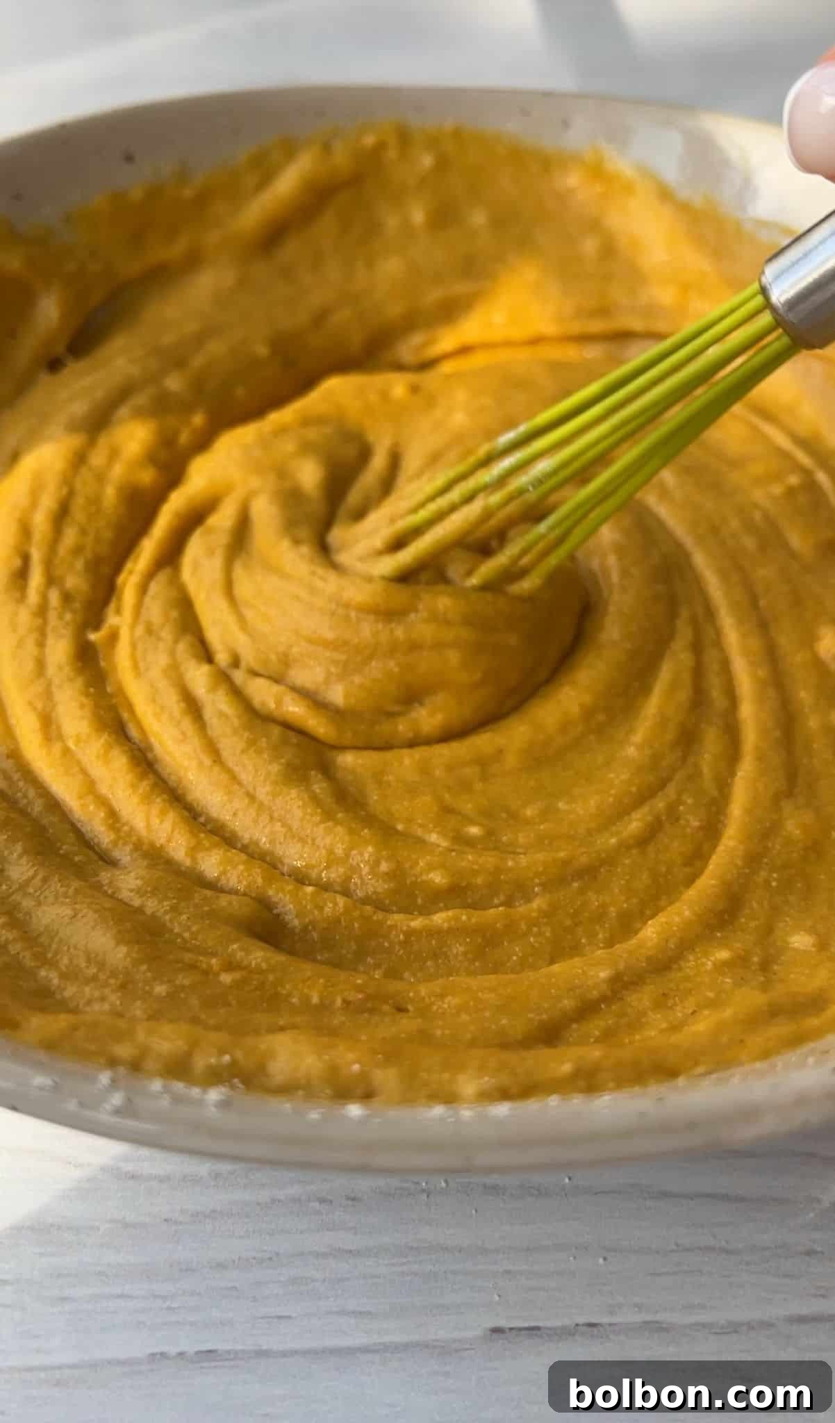 Wholesome Spiced Pumpkin Muffins 6 A close-up of a creamy, orange-hued mixture being whisked in a beige bowl. The texture appears smooth and thick. The bowl is placed on a light wooden surface.