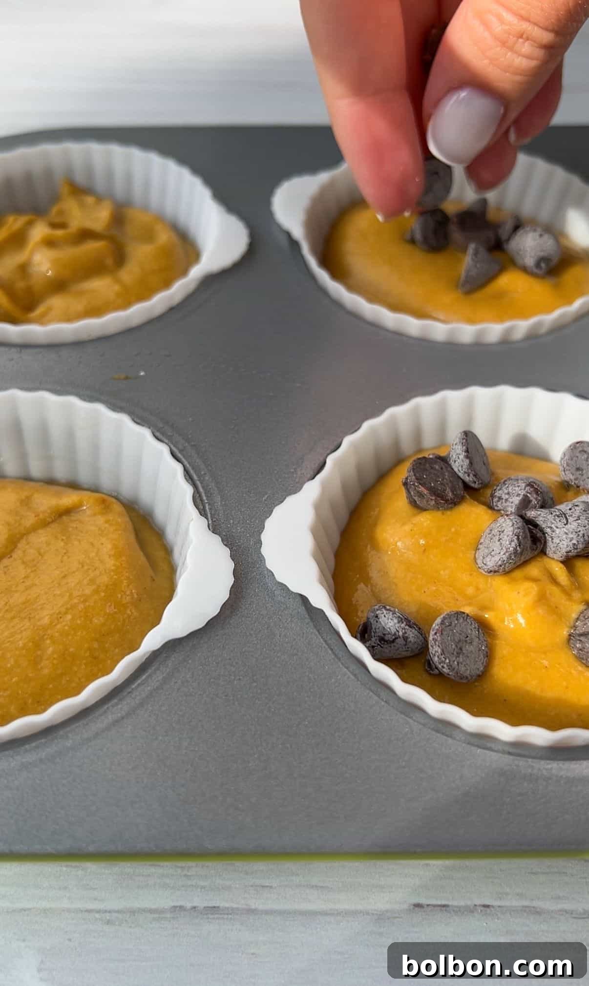 Wholesome Spiced Pumpkin Muffins 7 A hand sprinkles chocolate chips over batter in silicone muffin cups placed on a baking tray. The batter is a golden color, filling four cups, ready for baking.