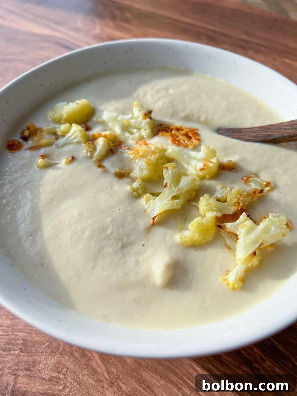 A bowl of creamy cauliflower soup garnished with roasted cauliflower florets. A wooden spoon rests in the bowl. The soup has a smooth texture, and the dish is placed on a wooden table surface.