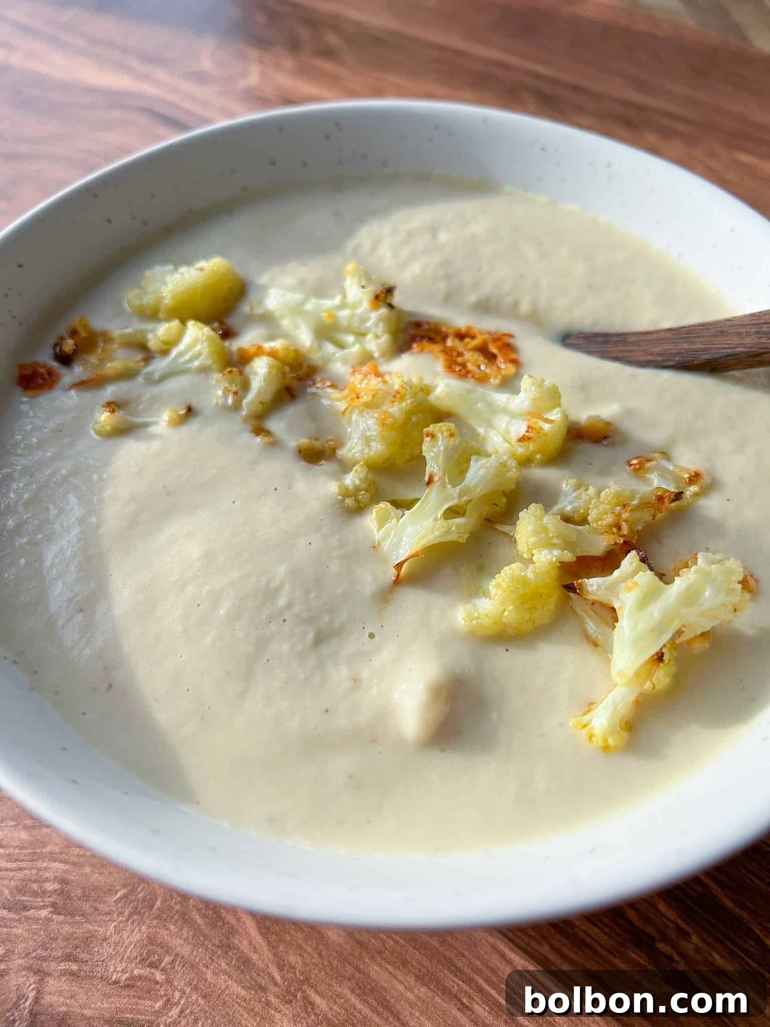 A bowl of creamy cauliflower soup garnished with roasted cauliflower florets. A wooden spoon rests in the bowl. The soup has a smooth texture, and the dish is placed on a wooden table surface.