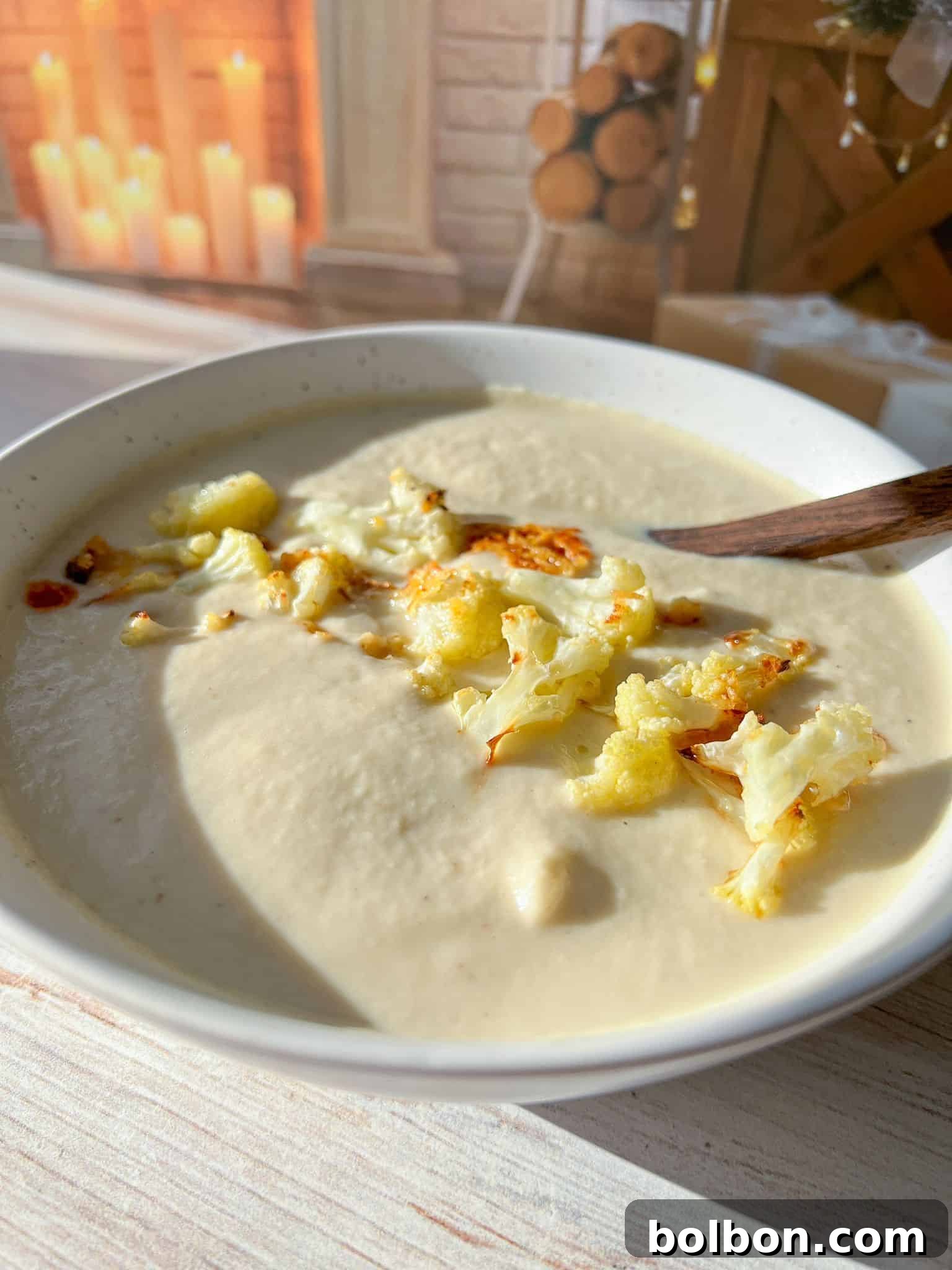 A bowl of creamy cauliflower soup topped with roasted cauliflower florets. A wooden spoon rests in the bowl. In the background, there's a cozy setting with candles and stacked firewood.