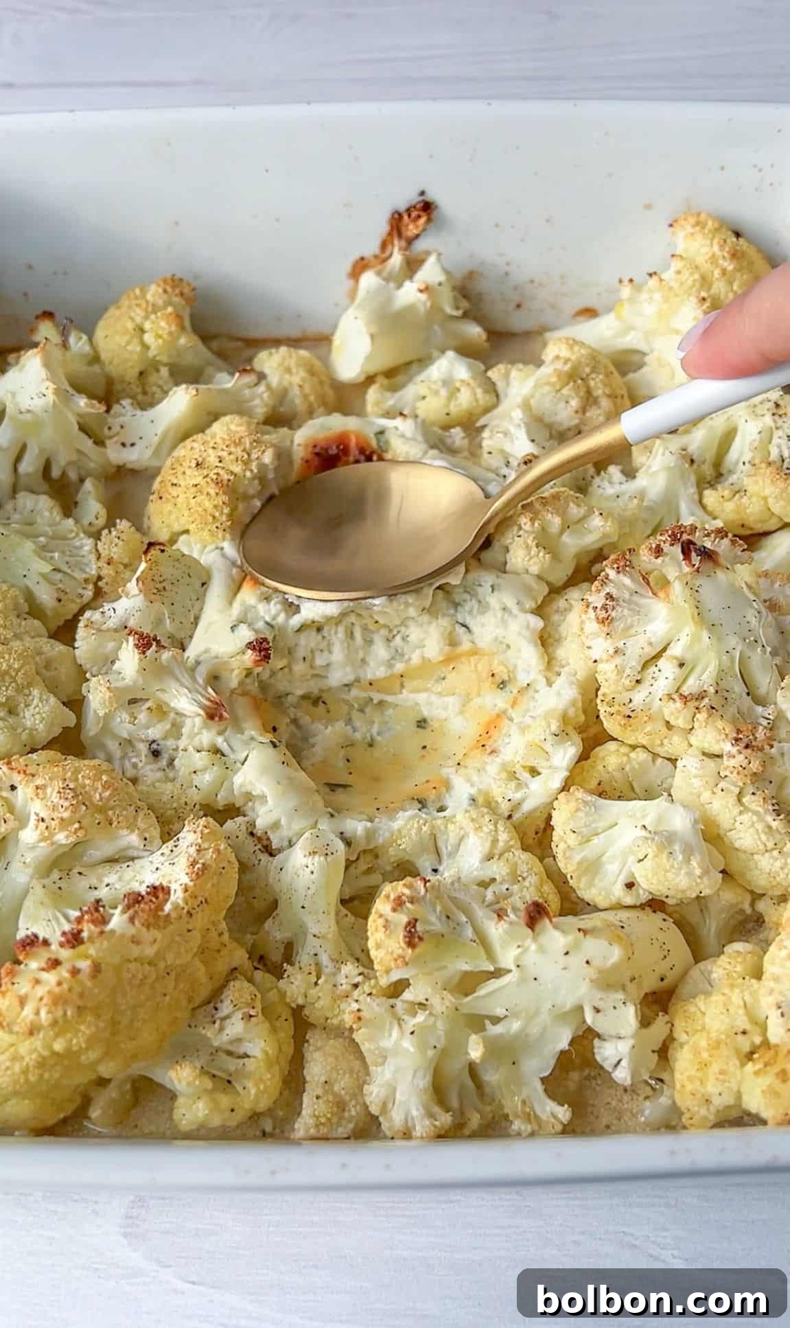 A hand holds a spoon, scooping creamy cheese sauce over roasted cauliflower florets in a white baking dish. The cauliflower is lightly browned and seasoned with herbs, creating a savory dish.