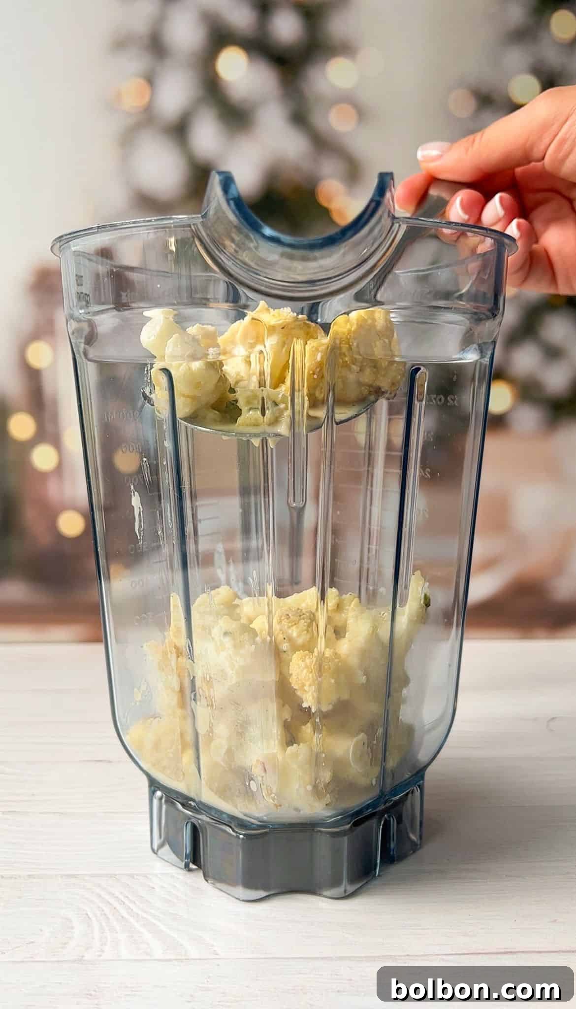 A blender jug with cauliflower pieces inside, held by a person. Blurry background with festive lights and trees.