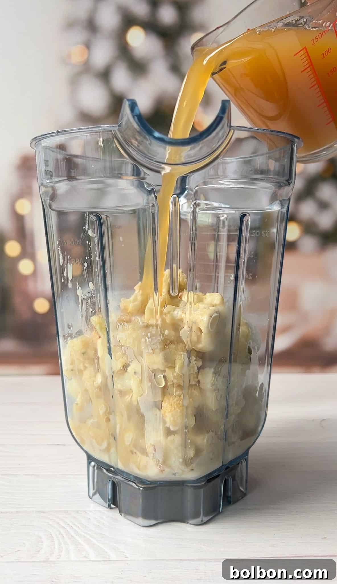 A clear blender jar filled with cooked pasta is placed on a white surface. Broth is being poured into the jar from a measuring cup. A blurred background shows warm holiday lights and a decorated tree.