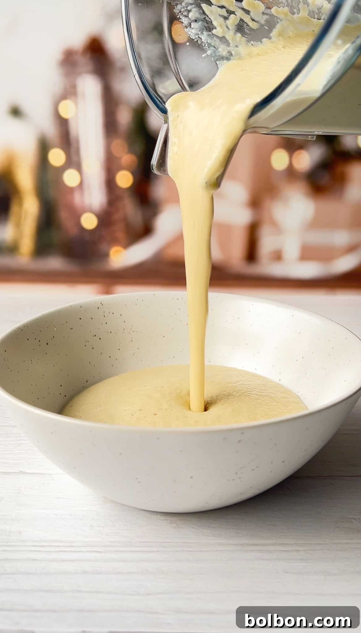 Pouring creamy yellow liquid from a blender jar into a speckled white bowl. The background is softly blurred, featuring warm lights and decorative elements. The scene suggests preparation for a recipe or meal.