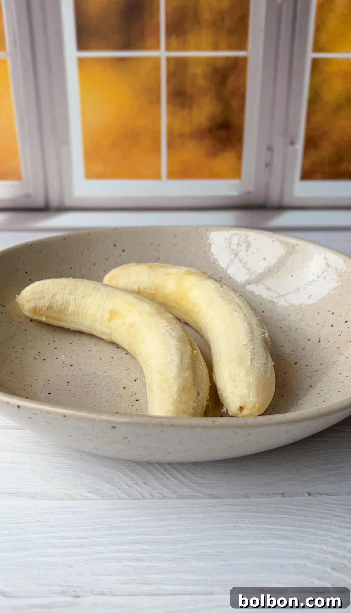 Two peeled bananas rest on a speckled ceramic plate. The plate is on a light-colored wooden surface, and in the background are windows showing a warm, blurred orange glow.