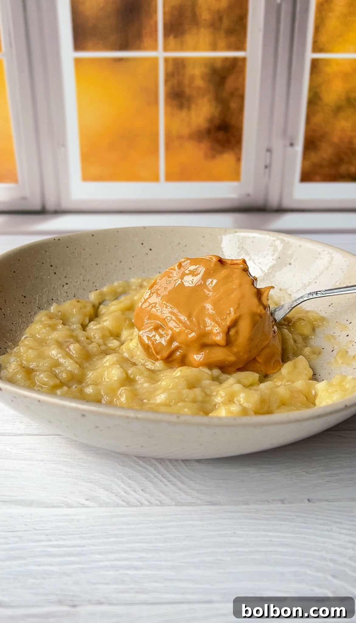 A bowl of mashed bananas with a spoonful of creamy peanut butter on top. The dish is set against a white wooden background with a window showcasing a warm, golden sunset view.