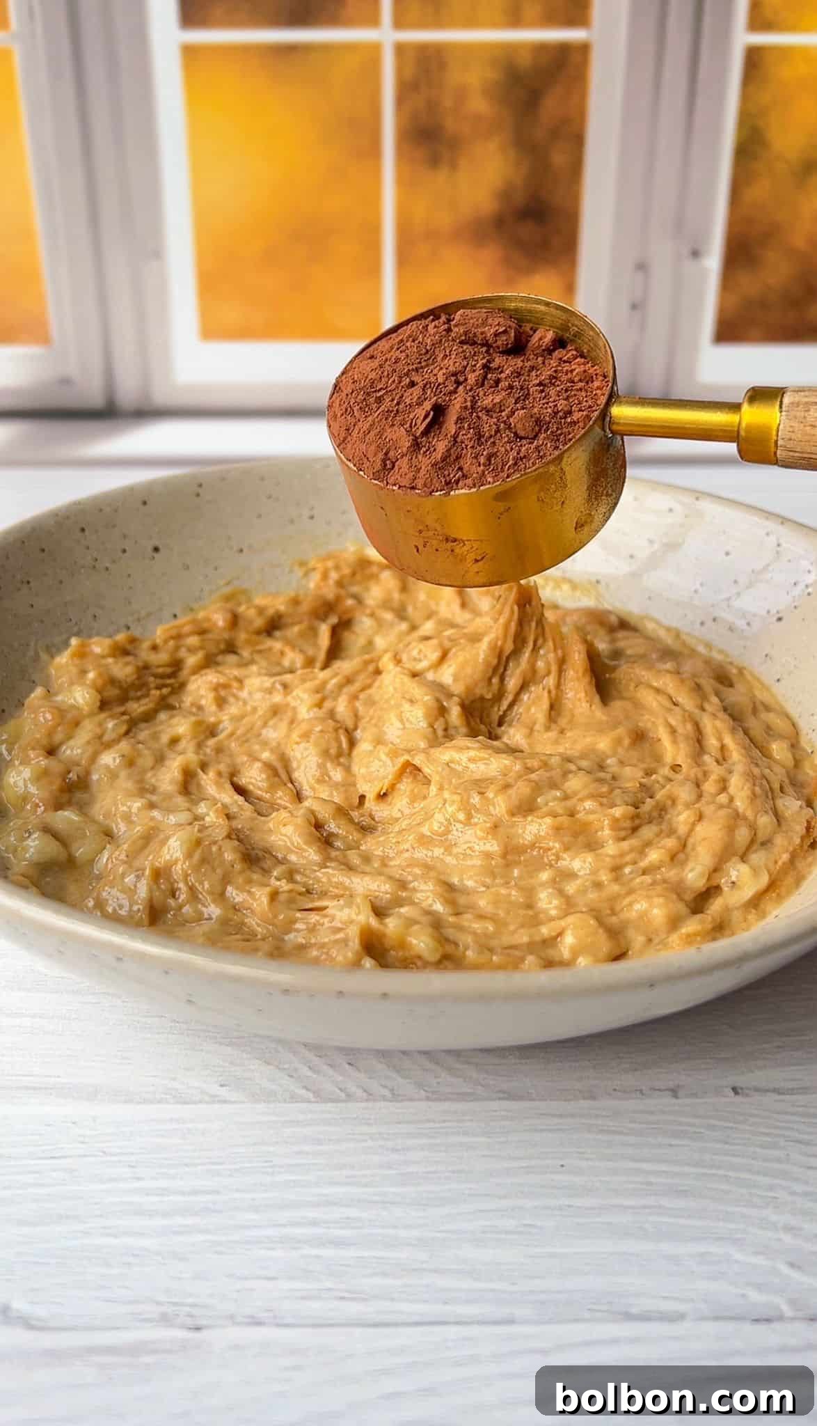 A bowl of mashed bananas sits on a white wooden table. Above the bowl, a golden measuring cup holds cocoa powder, ready to be added. The background features a window with a warm, sunny view.