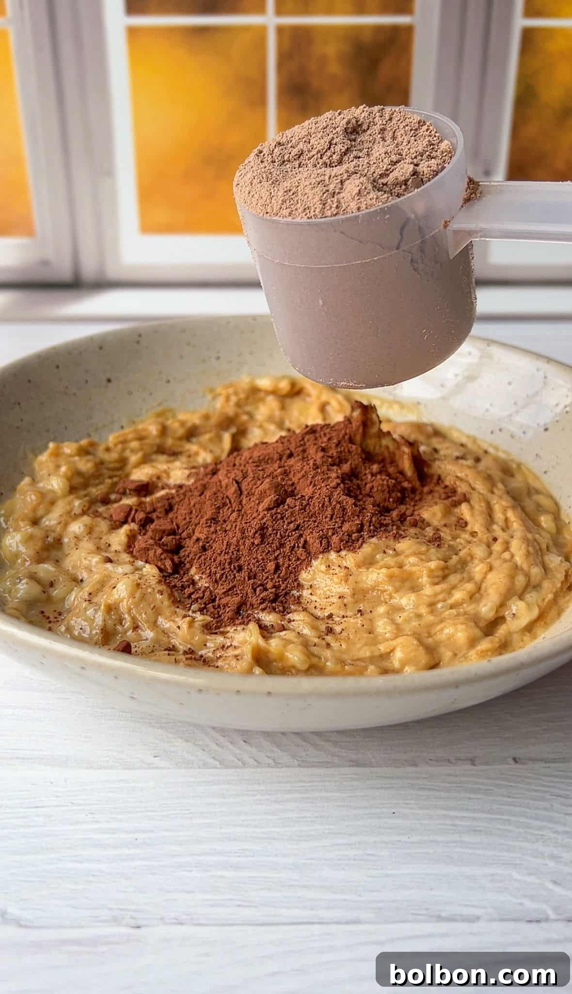 A bowl of oatmeal topped with a heap of cocoa powder. A hand holds a scoop of cocoa powder above the bowl, ready to add more. The background features a window with a warm, sunlit view.
