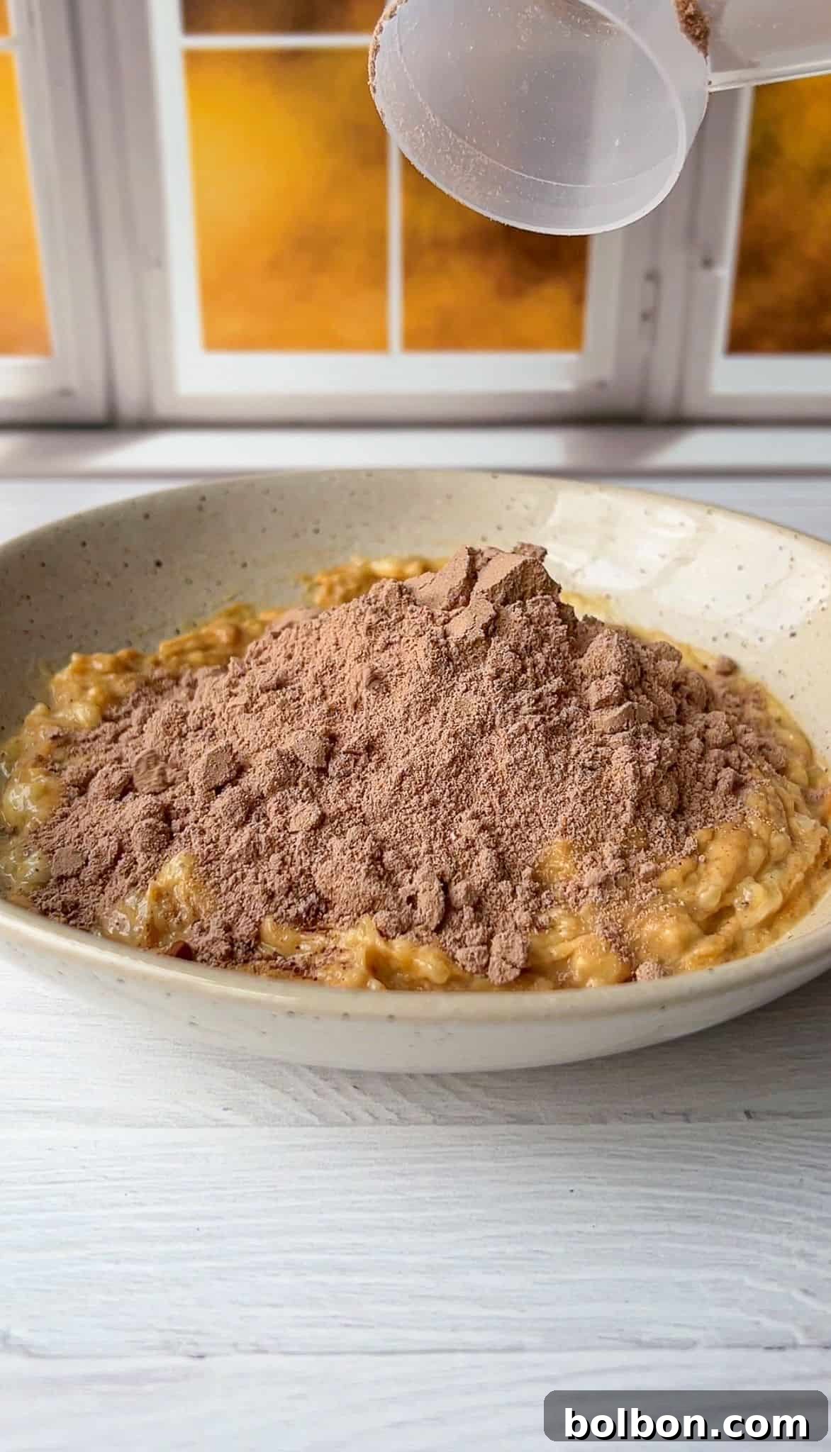 A bowl of oatmeal topped with a mound of cocoa powder on a light wooden table. A sifter is pouring more cocoa powder onto the oatmeal. The background features a window with a warm, golden light.