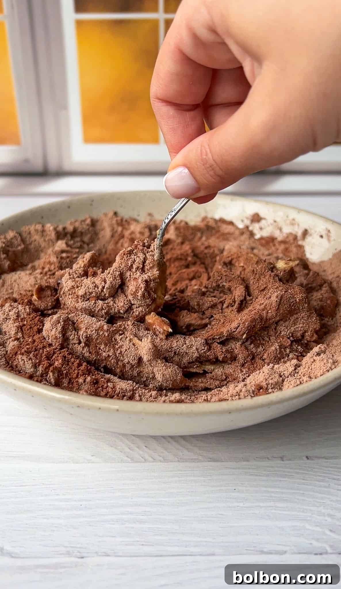 A hand holding a spoon mixes a bowl of cocoa powder and chopped nuts. The mixture is in a ceramic bowl on a light-colored wooden surface, with a blurred window in the background.