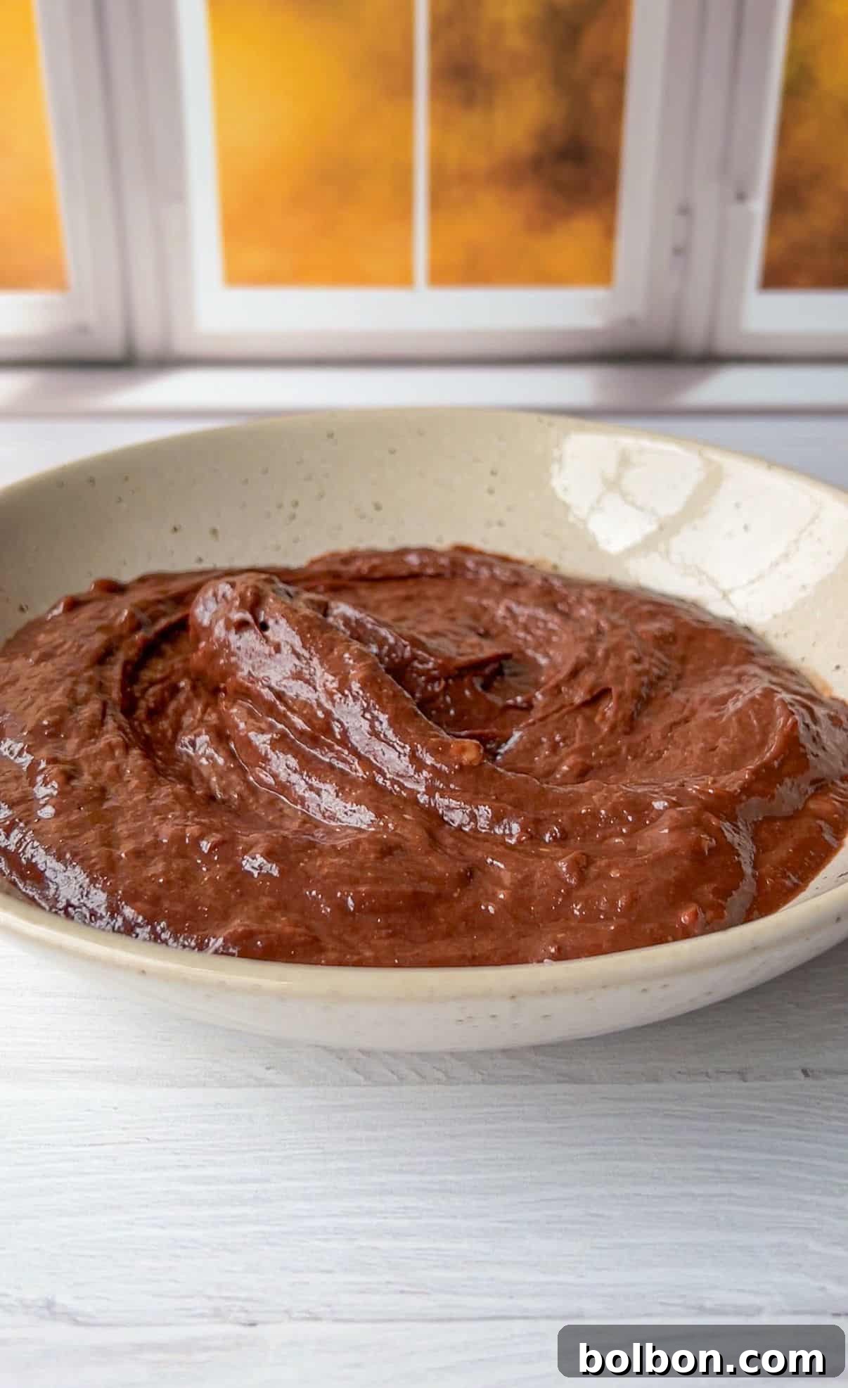 A bowl of thick, creamy chocolate pudding sits on a white wooden table. The smooth surface of the pudding glistens under natural light. A window with a warm glow is in the background.