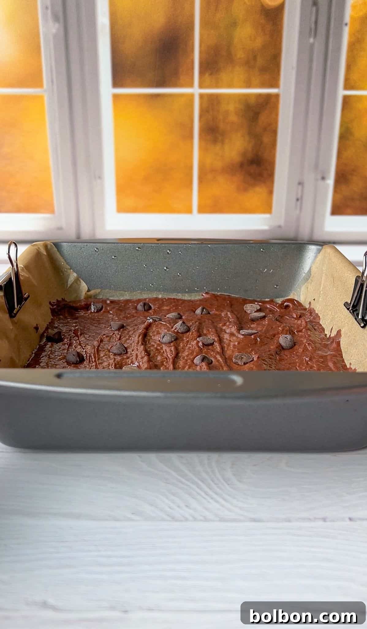 A baking pan with chocolate batter and chocolate chips on top is placed on a light-colored wooden surface. The pan is lined with parchment paper held by metal clips. A window with a soft orange glow is in the background.