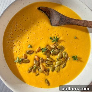 A bowl of creamy orange soup garnished with pumpkin seeds and small green herbs, served with a wooden spoon on a light wooden table.