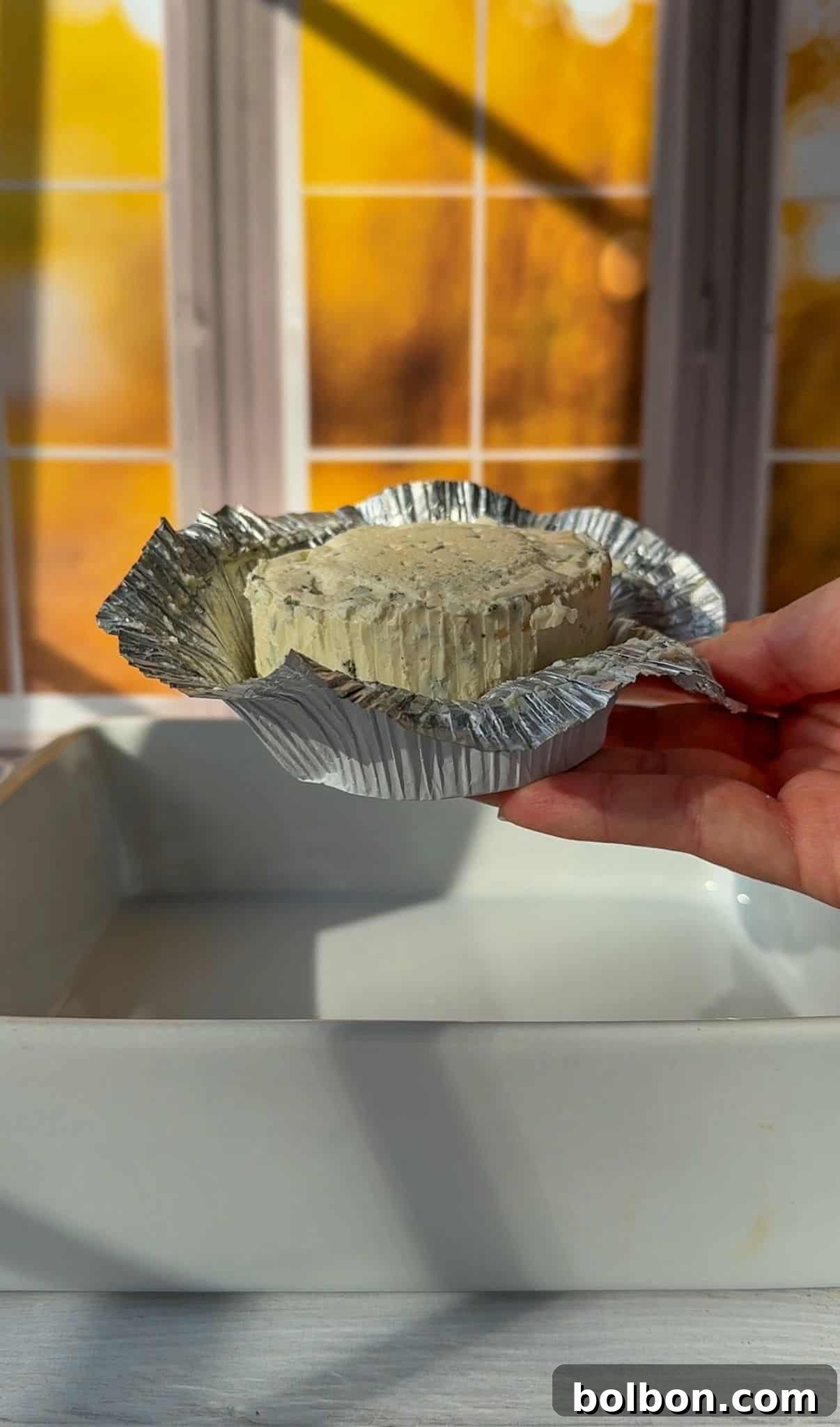 A hand holding an opened foil-wrapped round of cheese over a white dish. Warm sunlight streams through a window with a grid pattern in the background.