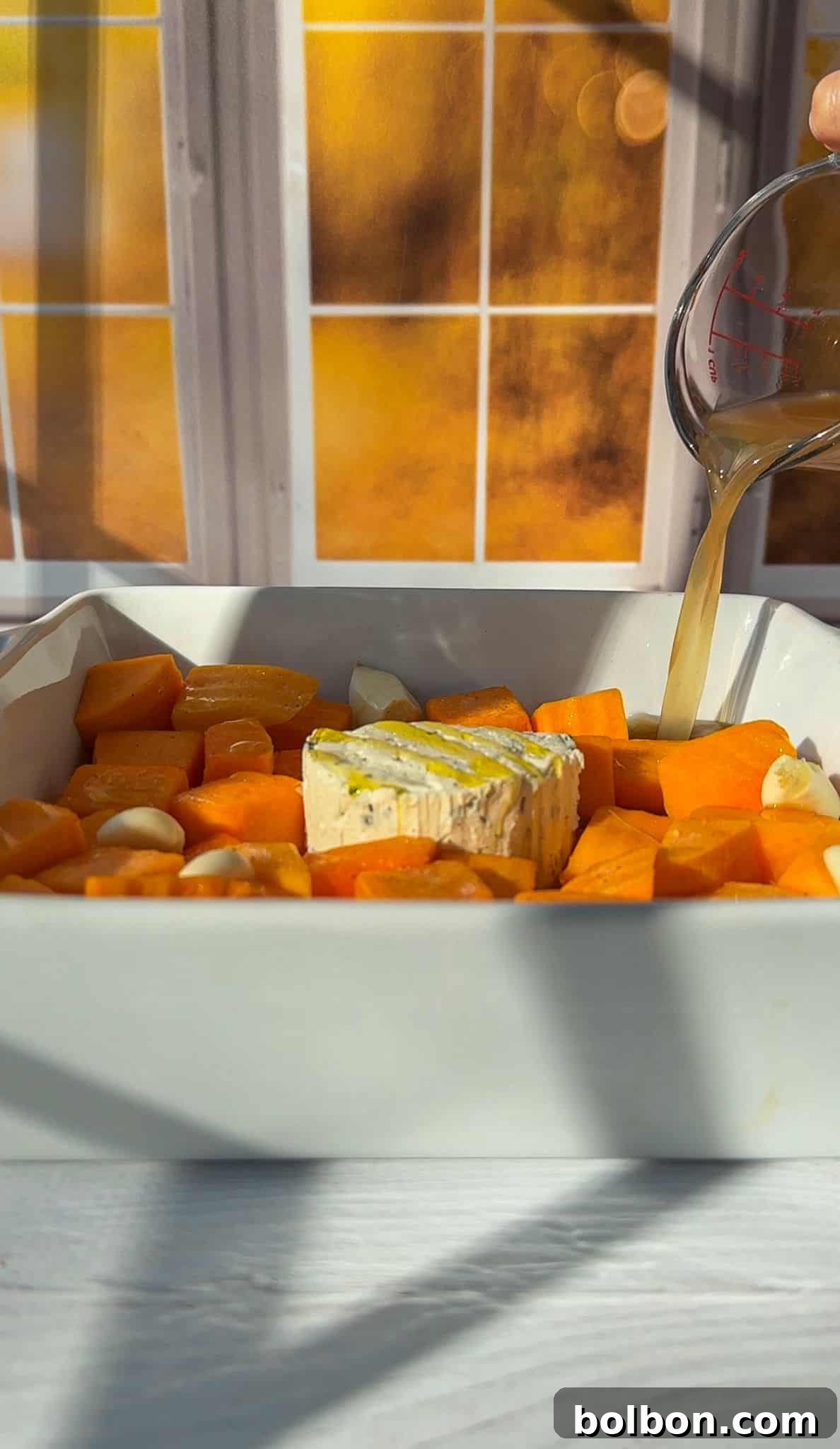 A baking dish filled with cubed sweet potatoes, garlic cloves, and a wheel of brie is shown. A hand is pouring broth from a measuring cup over the ingredients. A sunlit window with a warm orange glow is in the background.