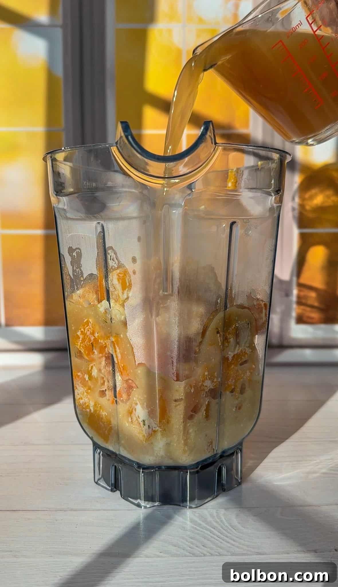 A blender container filled with squash, garlic and Boursin cheese is placed on a white countertop. A measuring cup is pouring a liquid into the blender. Sunlight filters through a window in the background, casting warm shadows.