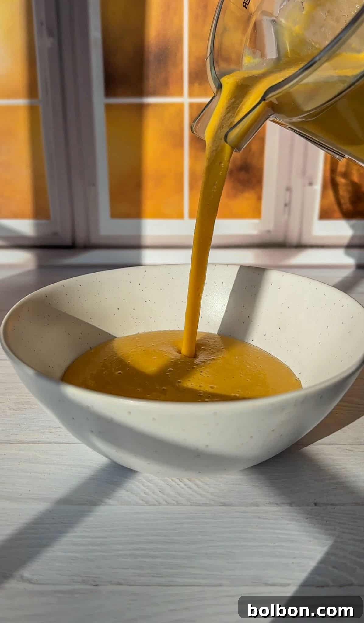 Smooth butternut squash soup being poured into a white speckled ceramic bowl, with a backdrop of sunlit windows.