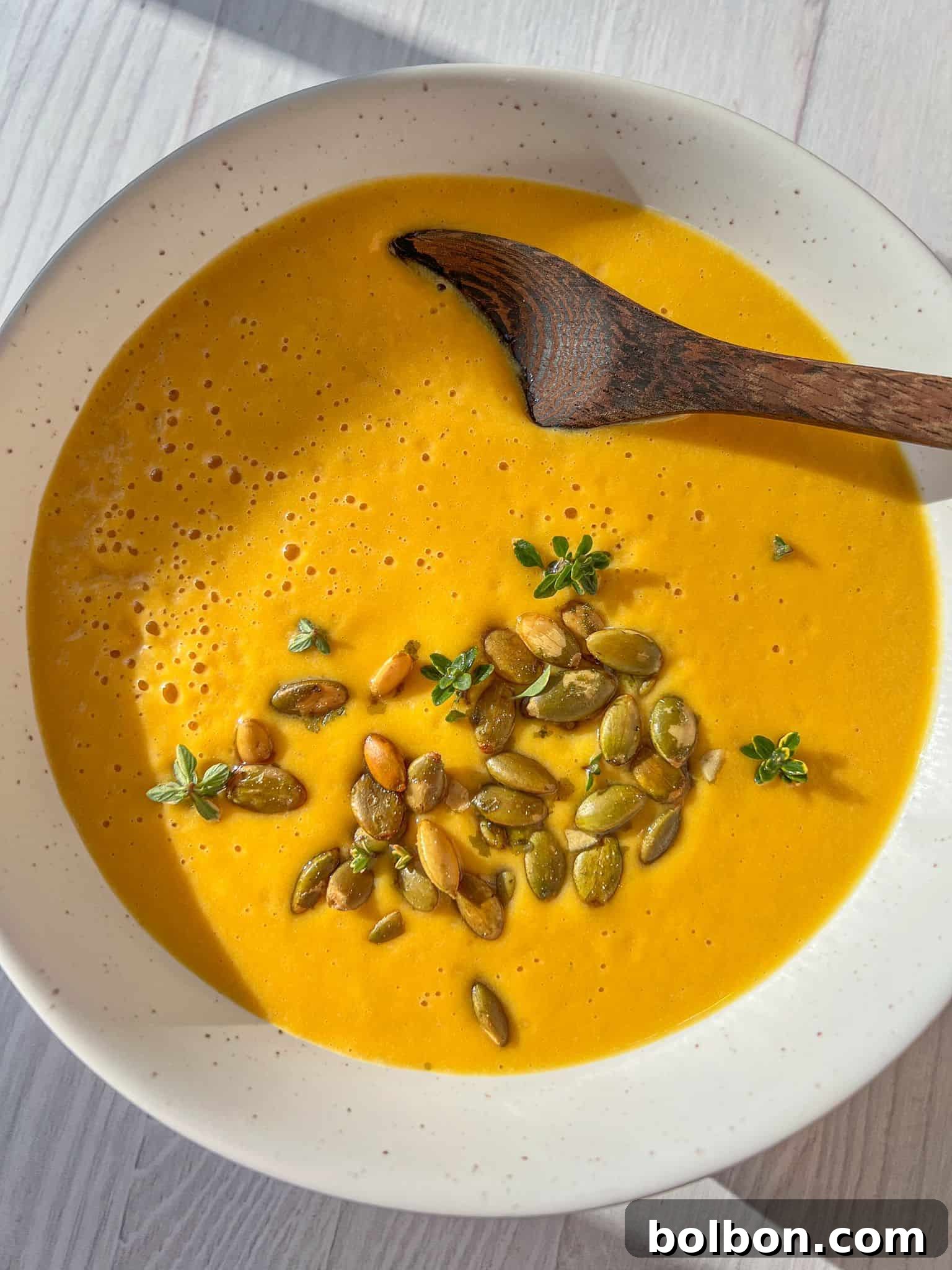 A bowl of creamy butternut squash soup garnished with pumpkin seeds and fresh thyme, with a wooden spoon resting inside. The soup is placed on a light-colored wooden surface, with sunlight casting soft shadows.