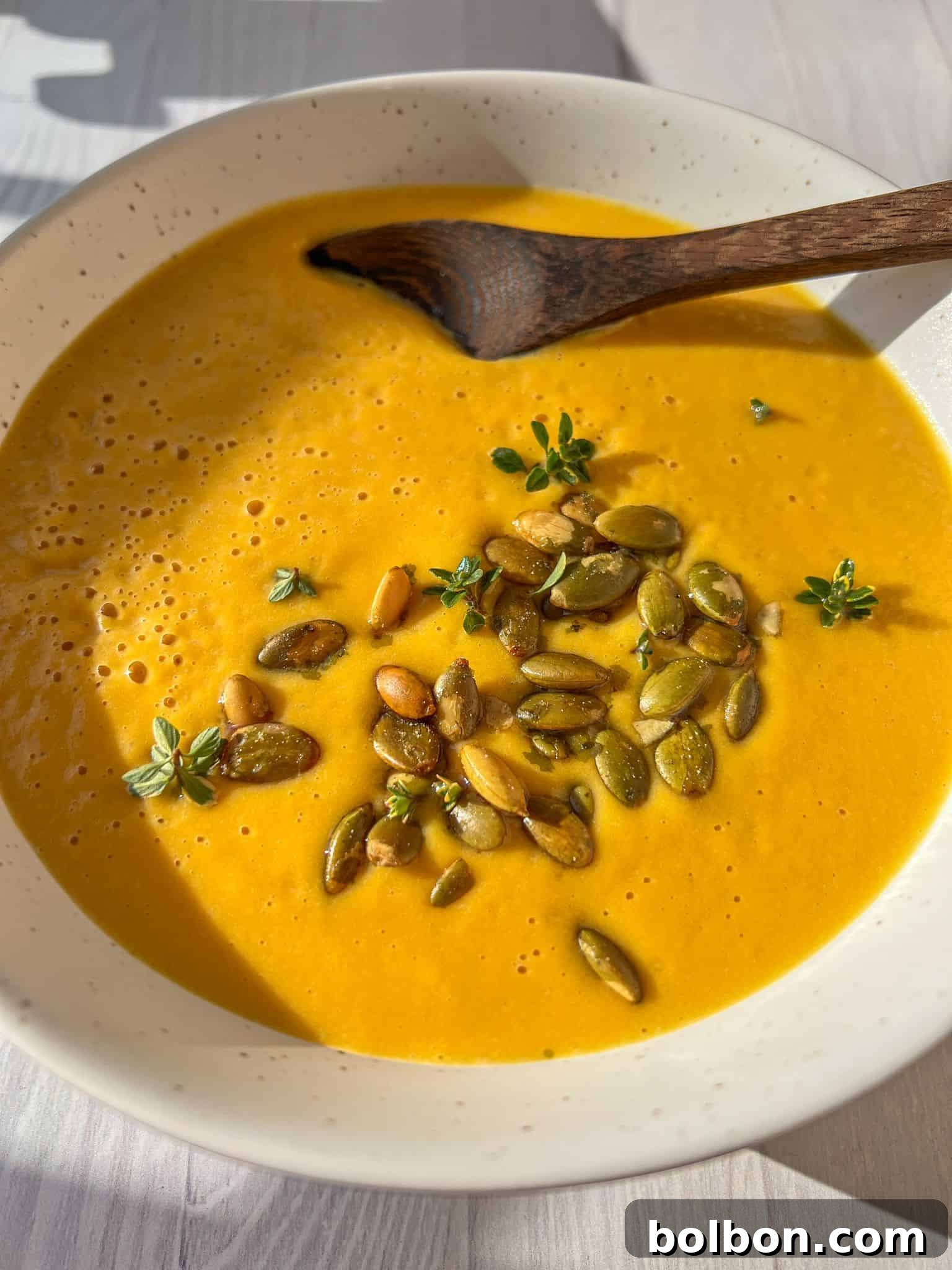 A creamy bowl of butternut squash soup garnished with pumpkin seeds and fresh thyme, served in a white bowl with a wooden spoon. Sunlight casts a natural shadow on the light-colored surface.
