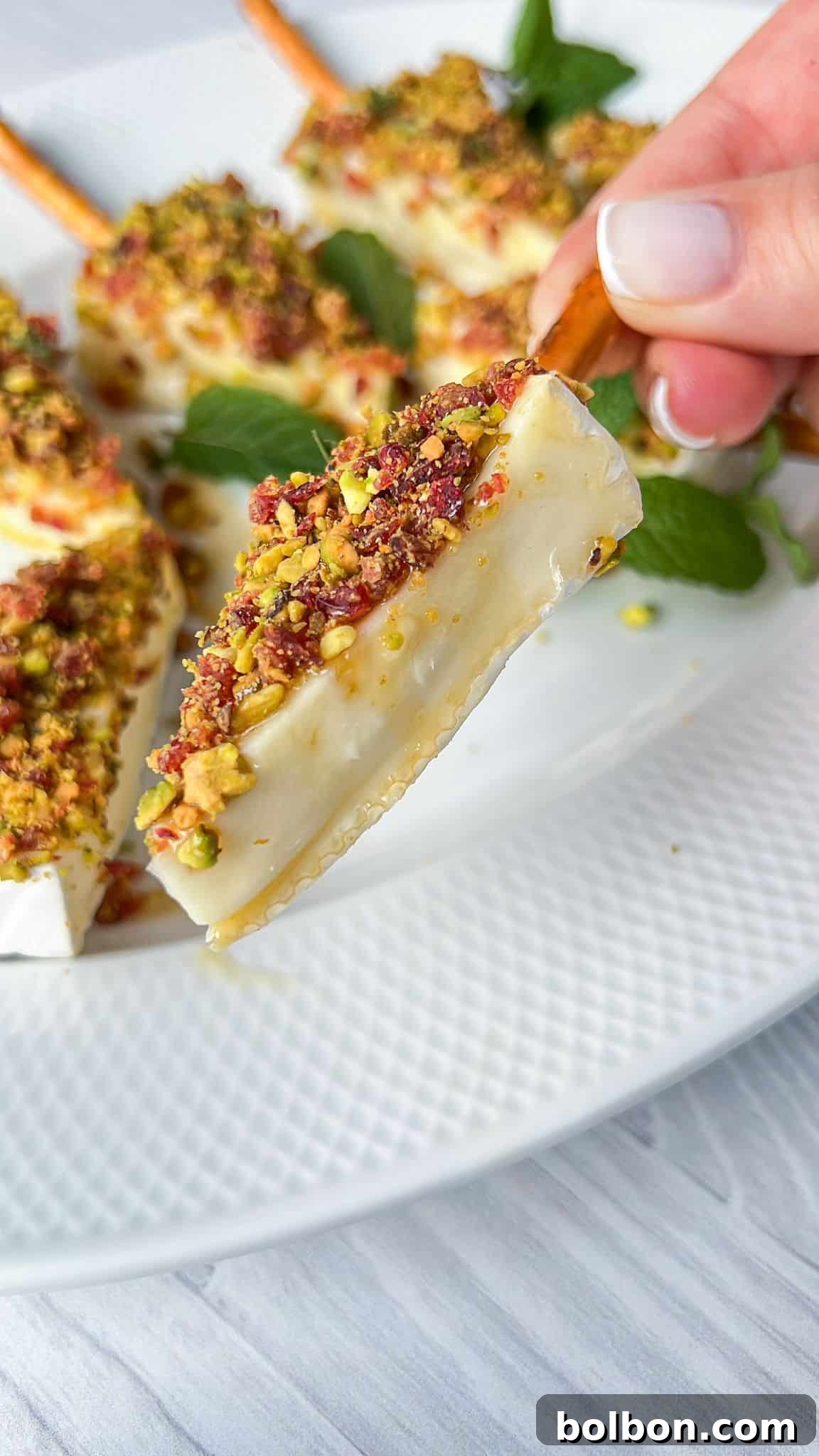 A close-up of a white plate featuring wedge-shaped holiday appetizers crusted with chopped nuts and herbs. A hand is seen holding one piece, highlighting its texture. Fresh mint leaves are used as garnish in the background.