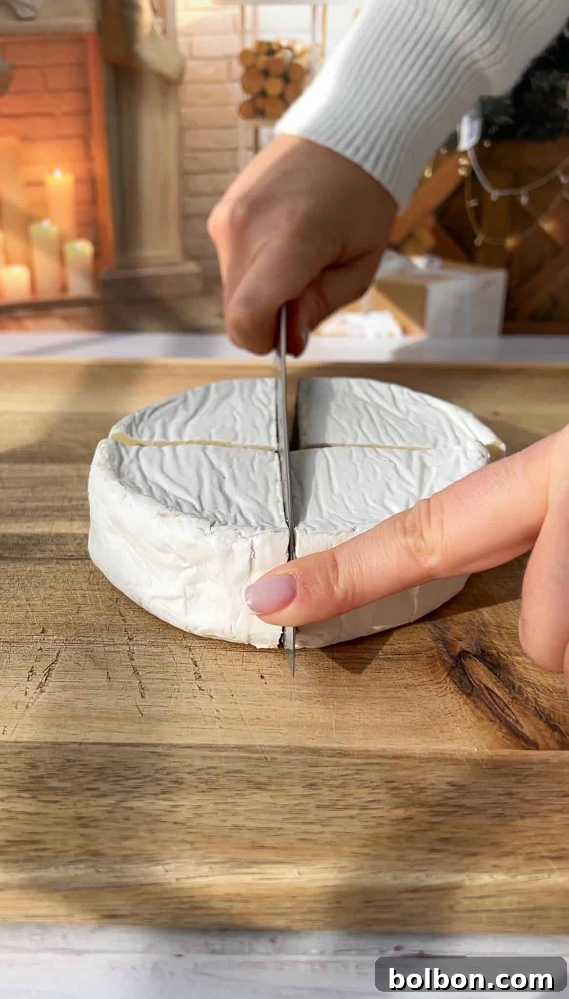 A person slicing a round wheel of brie cheese on a wooden cutting board. The background includes lit candles and stacked firewood, creating a warm and cozy atmosphere.