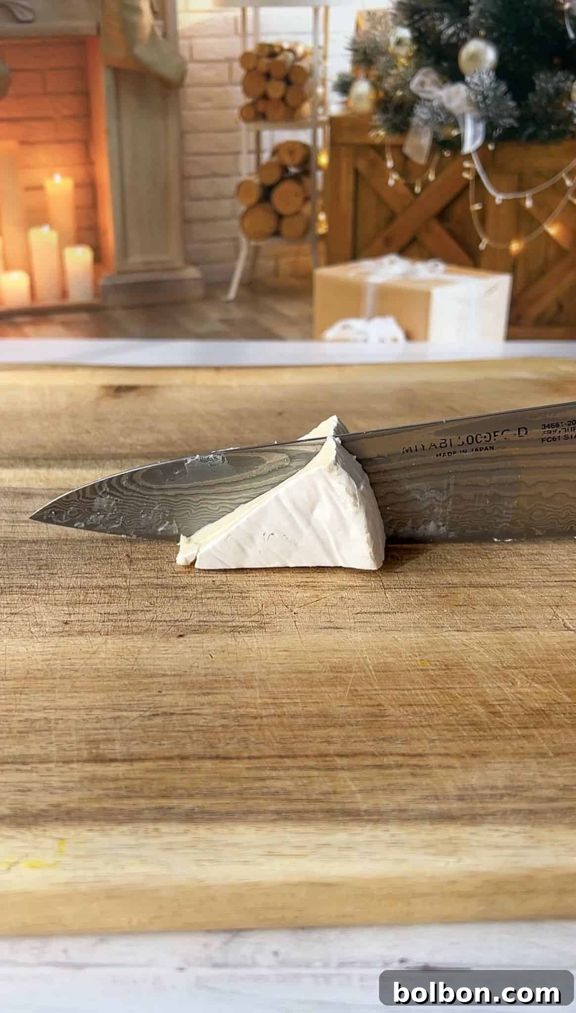 A knife slices through a piece of brie cheese on a wooden cutting board. The background features a cozy, festive room with candles, a fireplace, and holiday decorations.