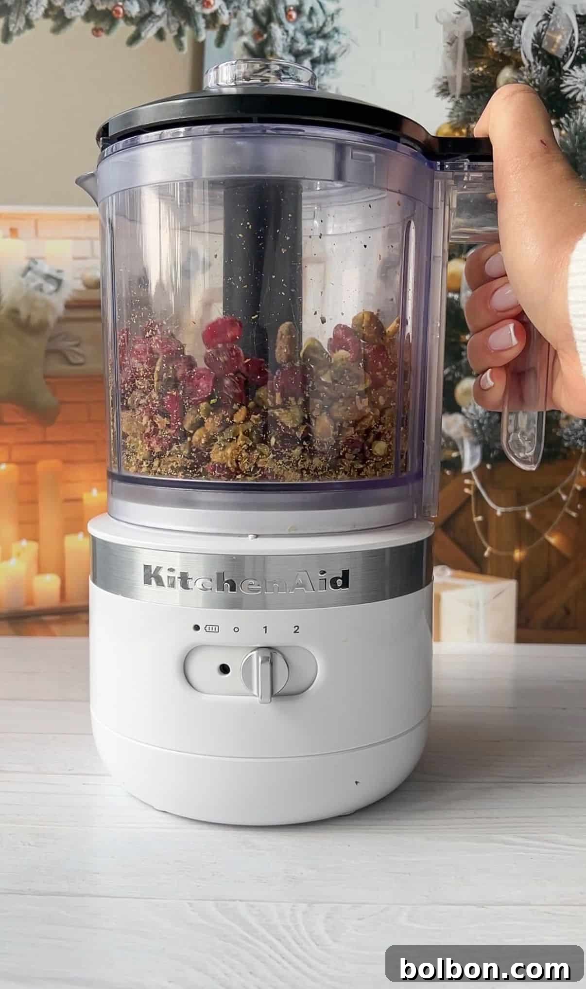 A hand holding the handle of a white KitchenAid food processor filled with dried fruits and nuts. The background shows a festive, decorated.