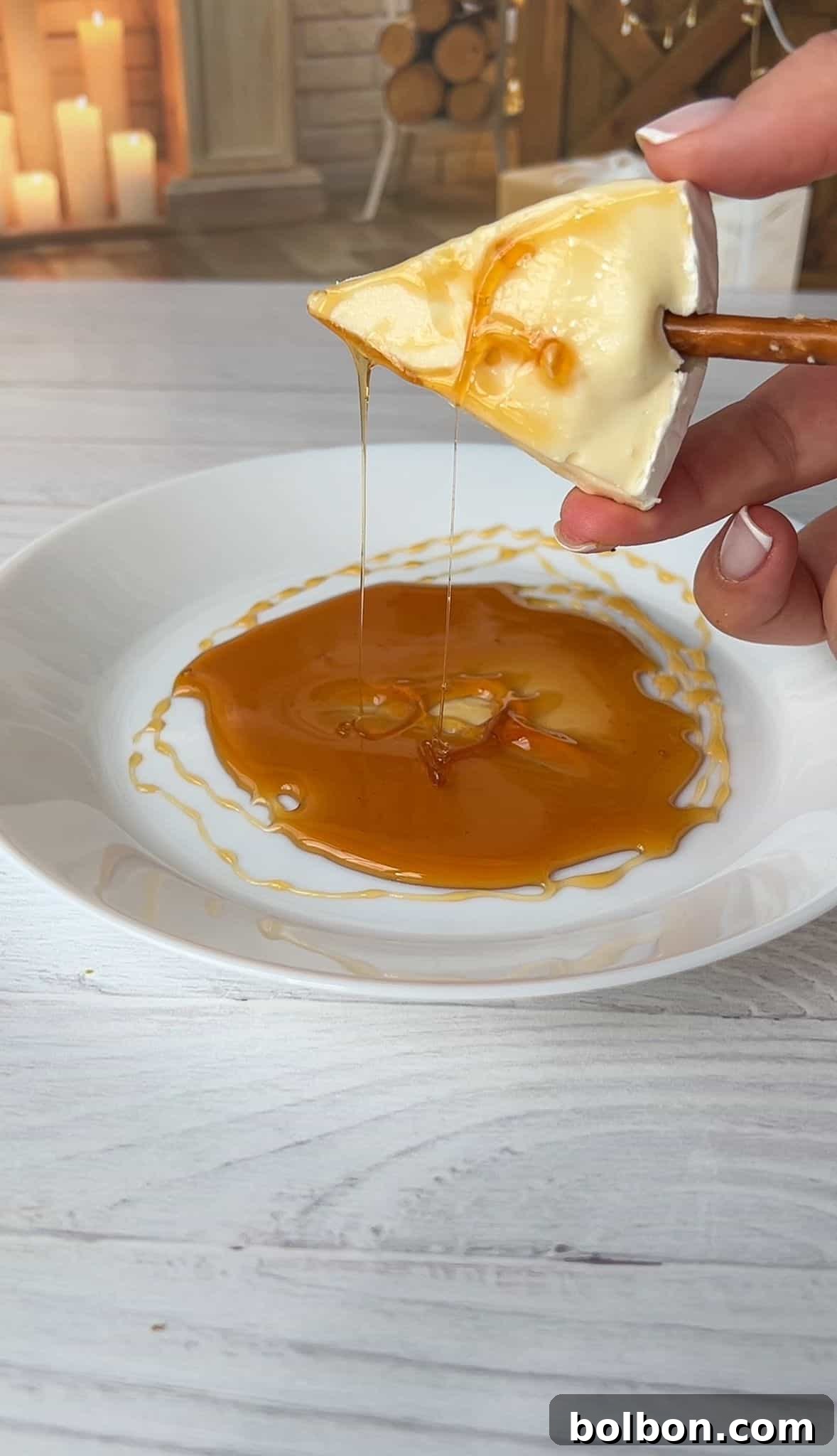 A piece of cheese is held above a white plate, dripping with honey. A pretzel stick is inserted into the cheese. The plate is on a white wooden surface with a cozy, candle-lit background.
