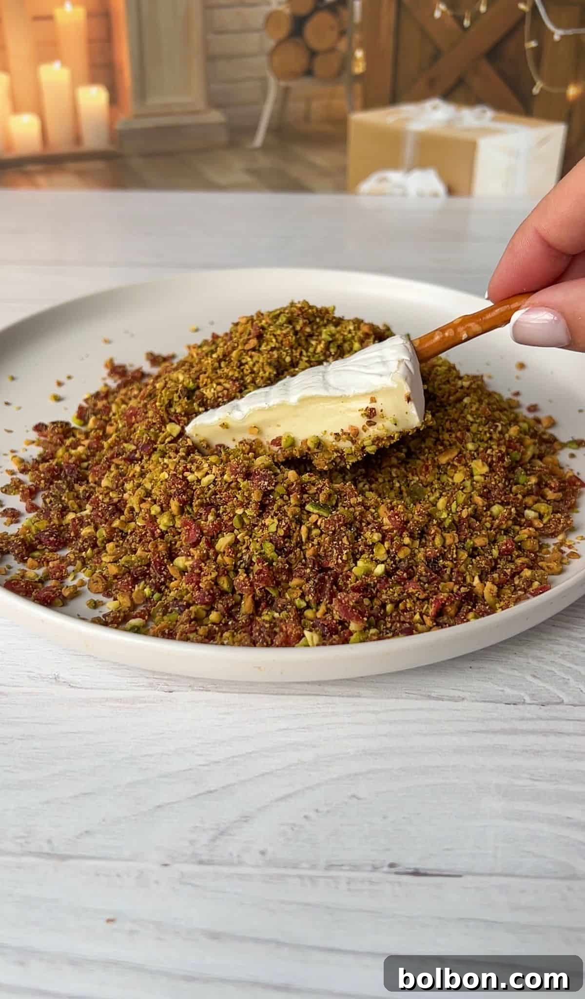 A wedge is being coated with a mixture of finely chopped nuts and herbs on a plate. In the background, there is a cozy setting with candles, logs, and a wrapped gift. 