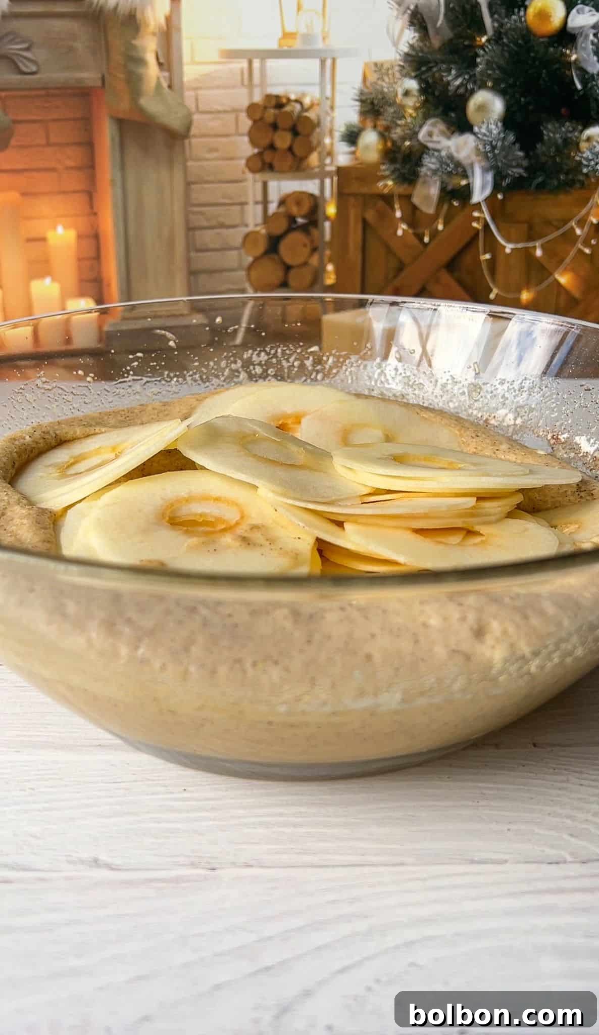 The Elusive Apple Cake 11 A large glass bowl filled with batter and thinly sliced apples is on a table. In the background, there's a cozy scene with a Christmas tree, a stack of firewood, and candles near a brick fireplace.