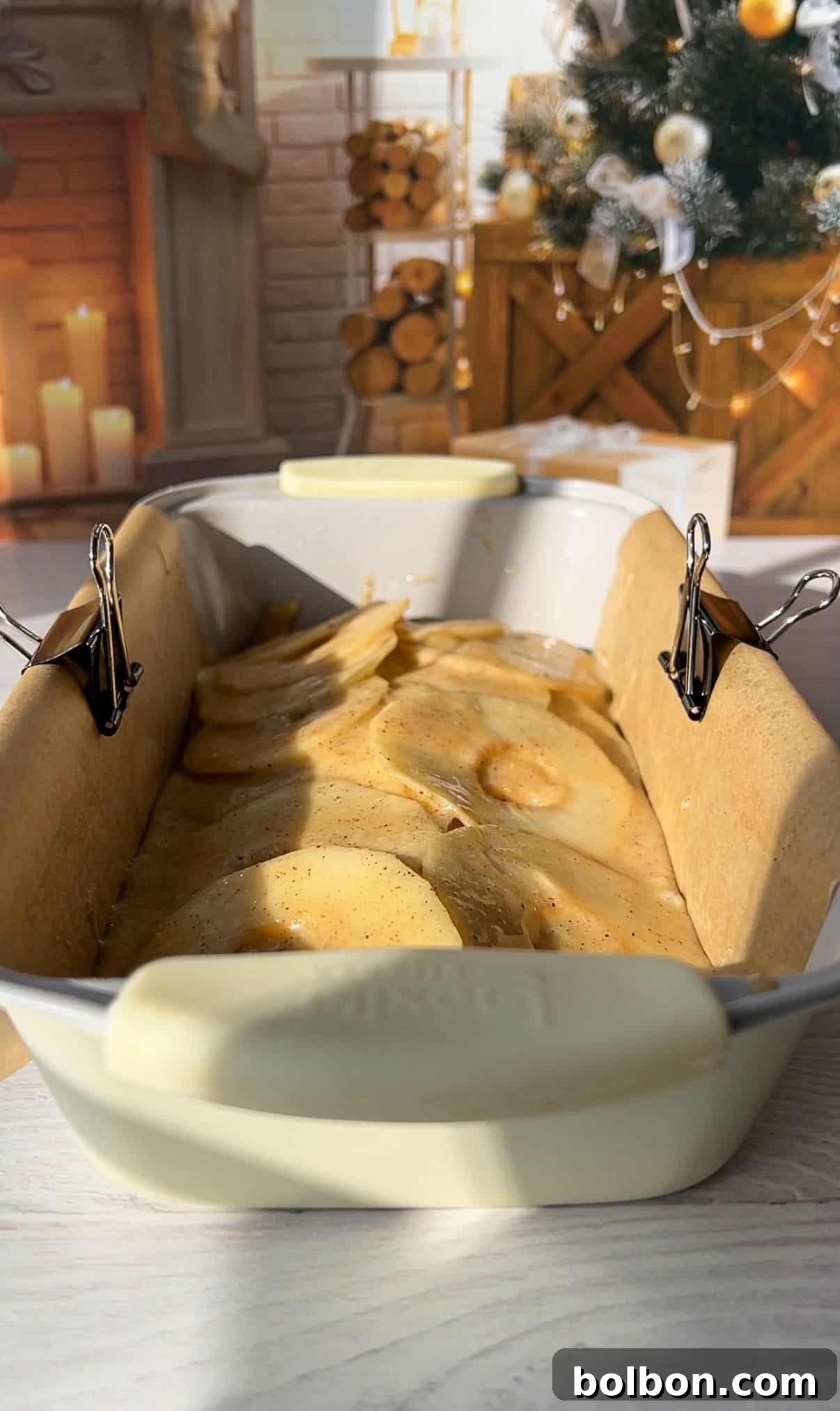 The Elusive Apple Cake 12 A loaf pan lined with parchment paper and secured with binder clips is filled with batter and apple slices. The background shows a cozy room with a decorated Christmas tree, a fireplace, stacked logs, and lit candles.