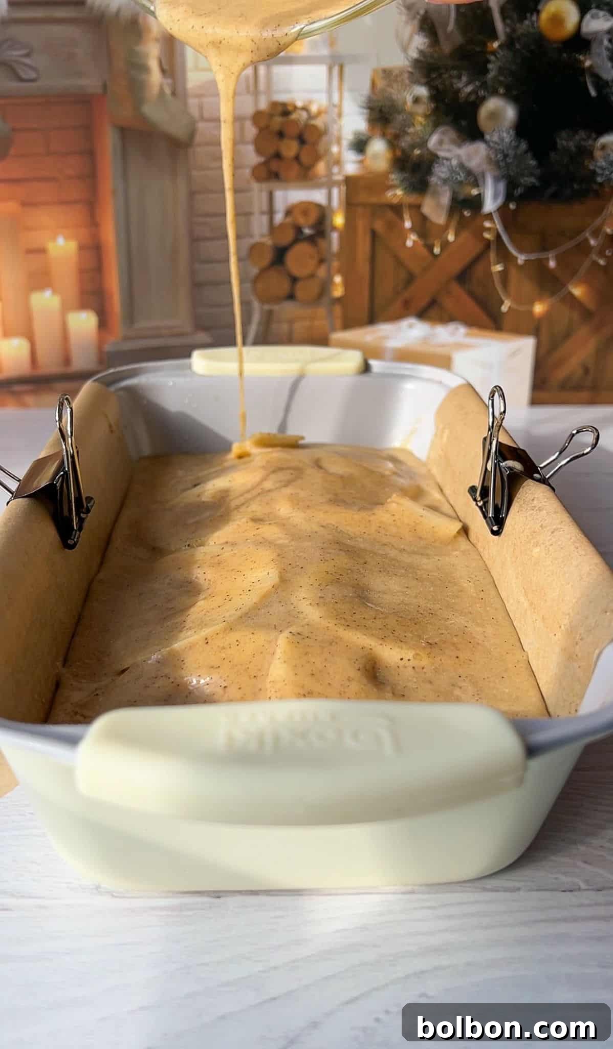 The Elusive Apple Cake 13 A creamy batter is being poured into a loaf pan lined with parchment paper, secured with binder clips. The background features a cozy fireplace and a decorated Christmas tree, creating a warm holiday atmosphere.