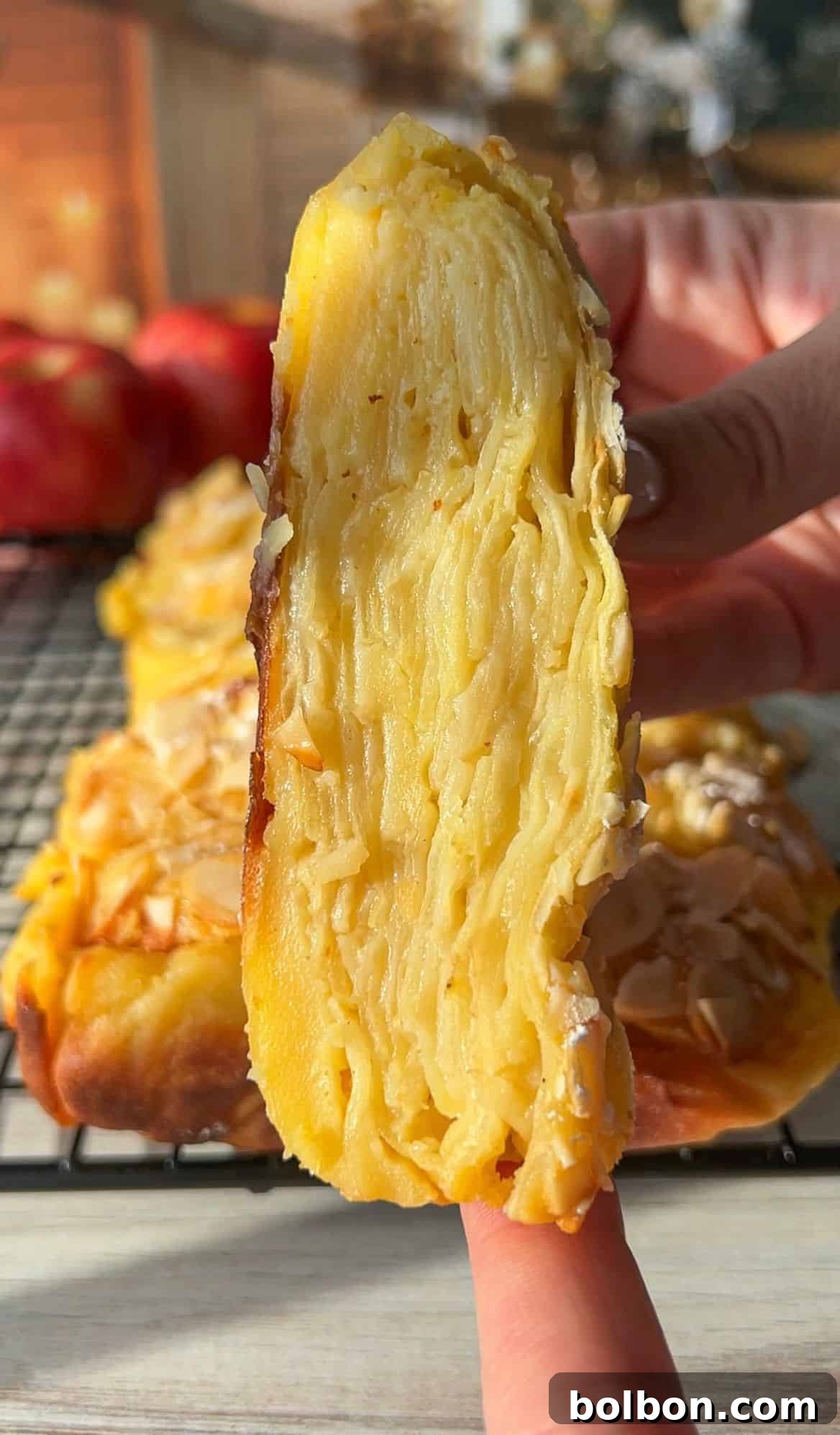The Elusive Apple Cake 14 Close-up of a hand holding a slice of invisible apple cake, showcasing the intricate layers within. The dessert has a golden crust, and sliced almonds are sprinkled on top. A cooling rack and red apples are visible in the blurred background.
