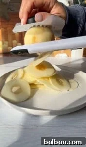 The Elusive Apple Cake 16 A hand uses a slicer to cut an apple into thin, uniform rings on a white plate. Sunlight casts shadows on the white wooden surface, and several sliced apple rings are neatly piled.