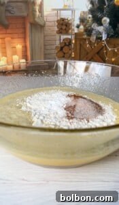 The Elusive Apple Cake 20 A glass bowl filled with cake batter and topped with flour and cocoa sits on a white kitchen counter. In the background, a decorated Christmas tree and a lit fireplace are visible, creating a cozy holiday atmosphere.