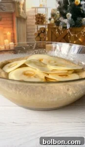 The Elusive Apple Cake 21 A large glass bowl filled with batter and thinly sliced apples is on a table. In the background, there's a cozy scene with a Christmas tree, a stack of firewood, and candles near a brick fireplace.