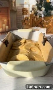 The Elusive Apple Cake 22 A loaf pan lined with parchment paper and secured with binder clips is filled with batter and apple slices. The background shows a cozy room with a decorated Christmas tree, a fireplace, stacked logs, and lit candles.