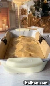 The Elusive Apple Cake 23 A creamy batter is being poured into a loaf pan lined with parchment paper, secured with binder clips. The background features a cozy fireplace and a decorated Christmas tree, creating a warm holiday atmosphere.