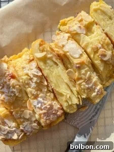 The Elusive Apple Cake 24 A close-up view of a sliced almond-topped pastry, dusted with powdered sugar. The golden layers of the pastry are visible, and a silver knife lies beside it on a wooden surface.
