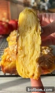 The Elusive Apple Cake 25 Close-up of a hand holding a slice of layered apple dessert, showcasing the intricate layers within. The dessert has a golden crust, and sliced almonds are sprinkled on top. A cooling rack and red apples are visible in the blurred background.