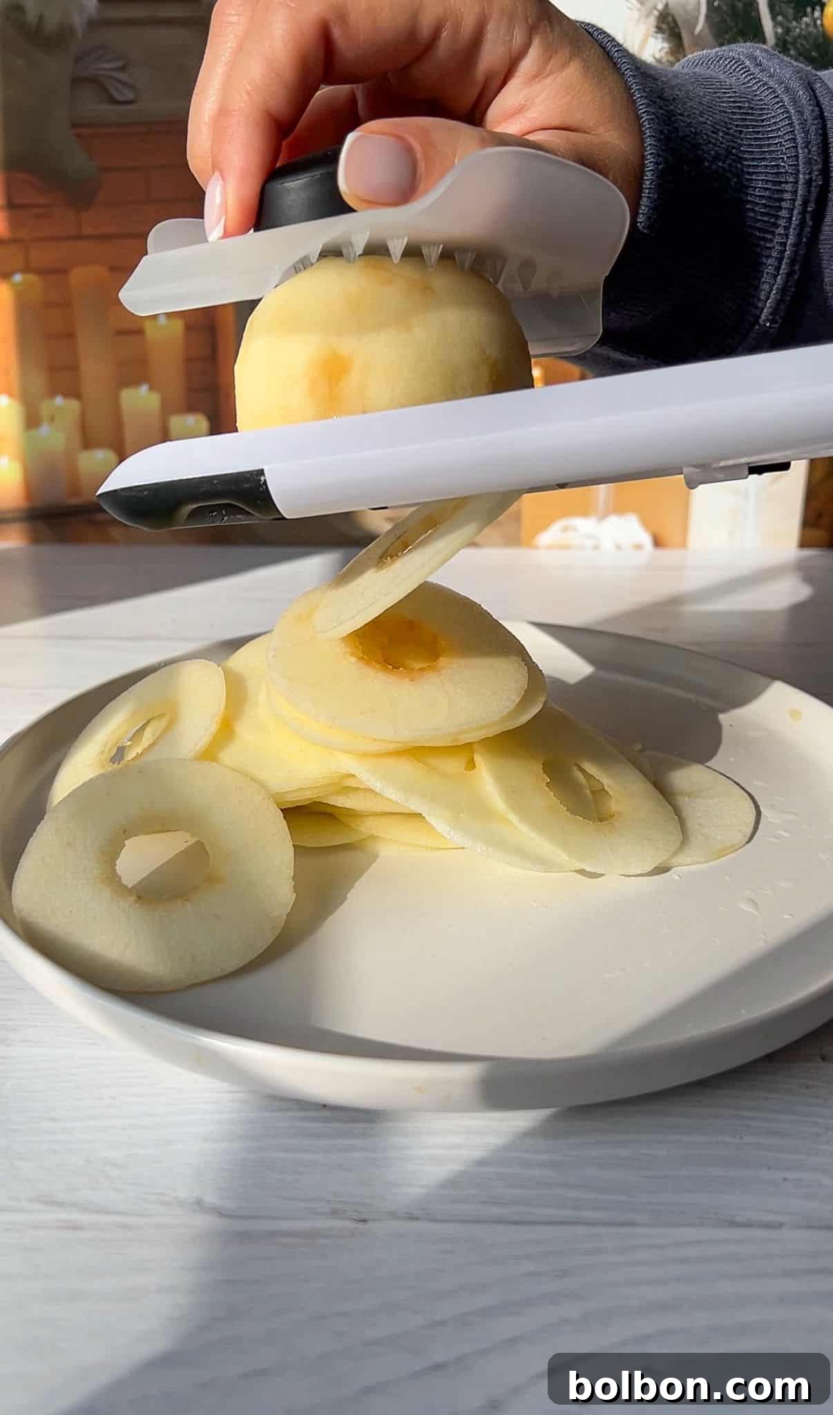 The Elusive Apple Cake 4 A hand uses a slicer to cut the fruit into thin, uniform rings on a white plate. Getting the apples prepared for the Invisible Apple Cake recipe.