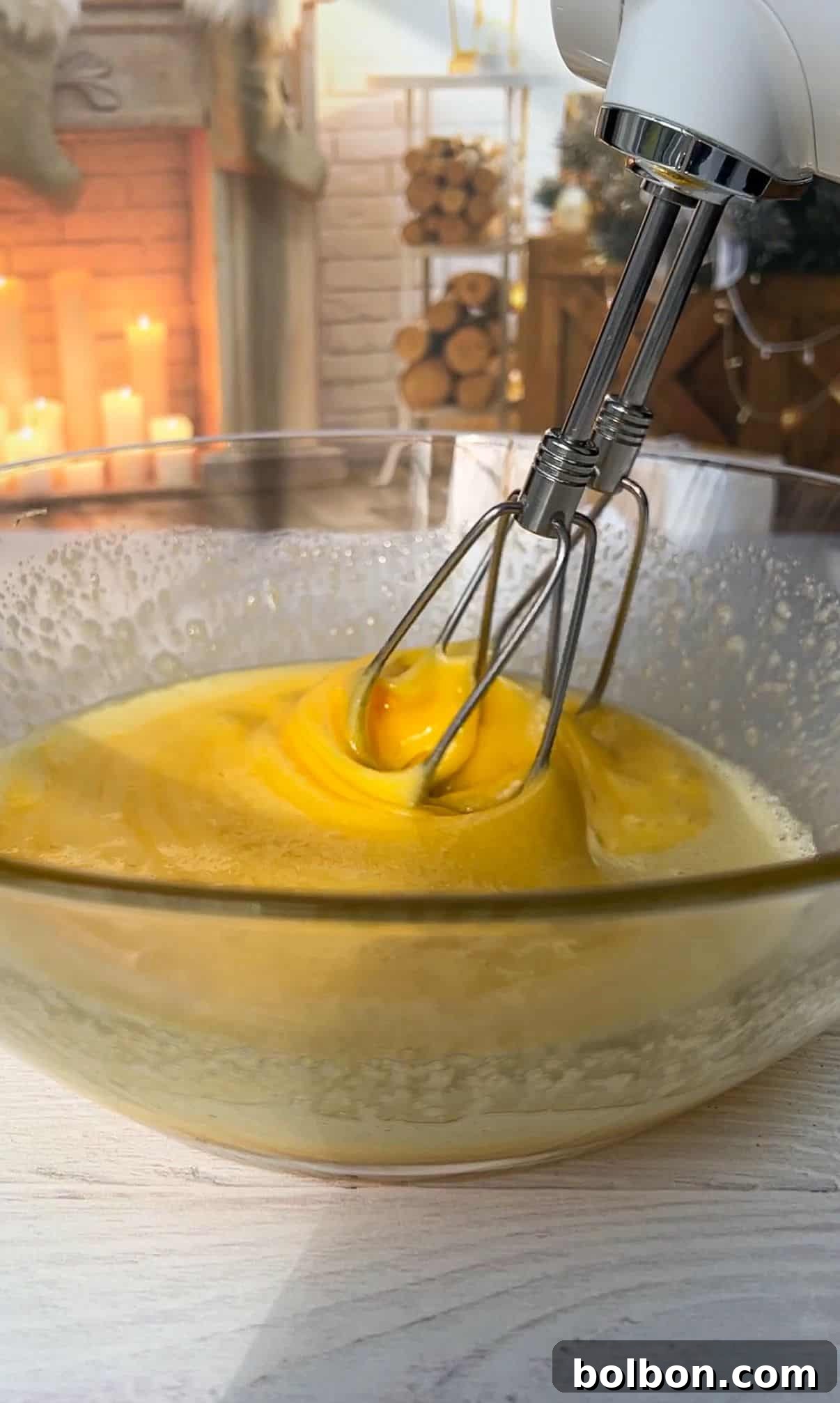 The Elusive Apple Cake 7 Close-up of an electric mixer beating eggs in a glass bowl. In the background, a cozy room with lit candles, stacked firewood, and a fireplace creates a warm ambiance.